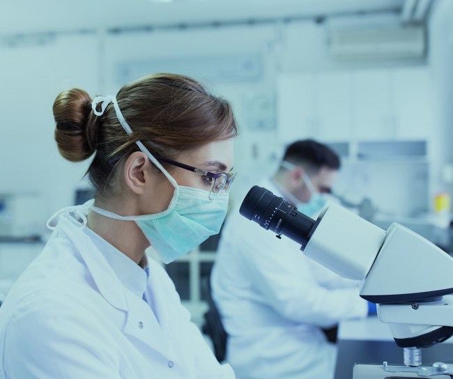 Eine Wissenschaftlerin mit Mundschutz und Schutzbrille sieht in einem modernen Labor durchs Mikroskop