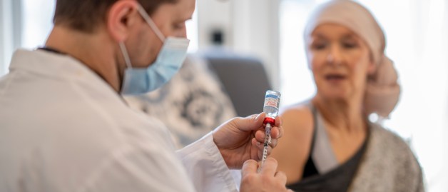 Ein Arzt mit Mundschutz zieht eine Spritze auf, Auf der Behandlungsliege sitzt eine Krebspatientin und schaut ihm zu