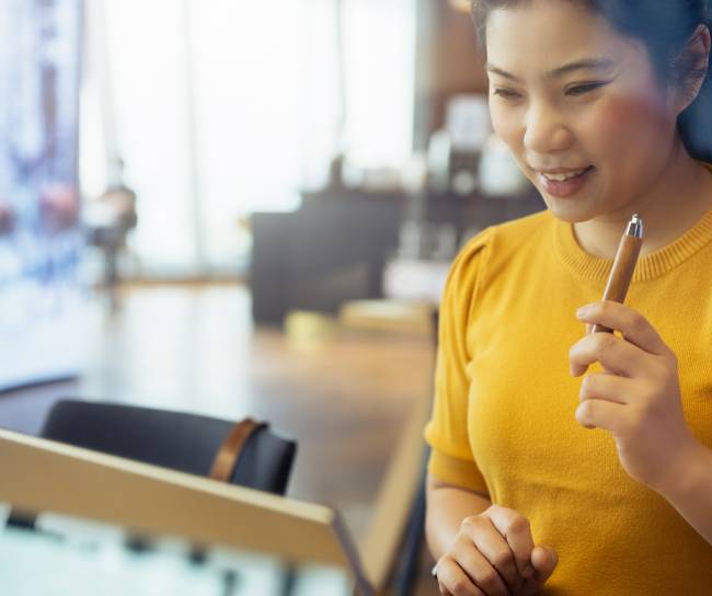 Eine Frau asiatischer Herkunft spricht interessiert in ein Tablet, welches vor ihr auf dem Tisch steht.