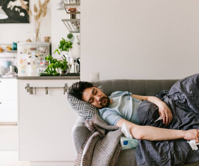 Ein kranker Mann mit Taschentuch in der Hand liegt auf dem Sofa unter einer Decke.