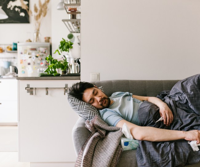 Ein kranker Mann mit Taschentuch in der Hand liegt auf dem Sofa unter einer Decke.