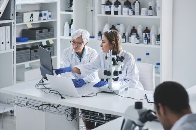 Zwei Labormitarbeiterinnen sitzen am Laptop und sprechen miteinander.