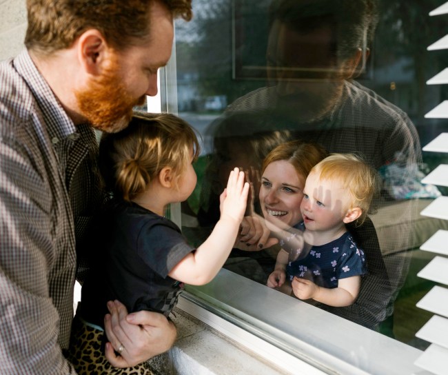 Ein Mann mit Kind auf dem Arm kommuniziert mit einer Frau mit Kind auf dem Arm durch eine Fensterscheibe. Beide Kinder halten die Hand an die Scheibe