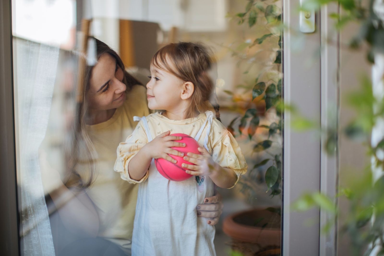 Eine Mutter und ihre kleine Tochter stehen hinter einem Fenster und sehen sich glücklich an.