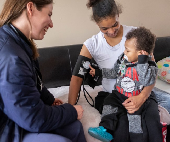 Zwei Frauen sitzen auf dem Sofa. Die rechte Frau hat eine Blutdruckmanschette um den rechten Arm und ein Kind auf dem Schoß.