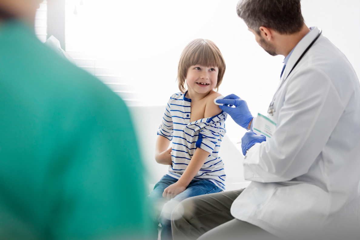 Kleiner Junge sitzt auf der Liege beim Arzt und lacht diesen an. Der Arzt ist zum Kind gewandt und desinfiziert mit einem Wattepad den Oberarm