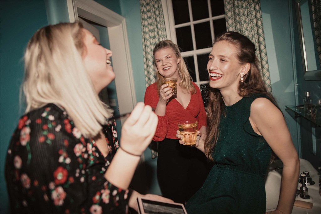 Drei Freundinnen stehen lachend in einer Altbauwohnung, Drinks in den Händen.