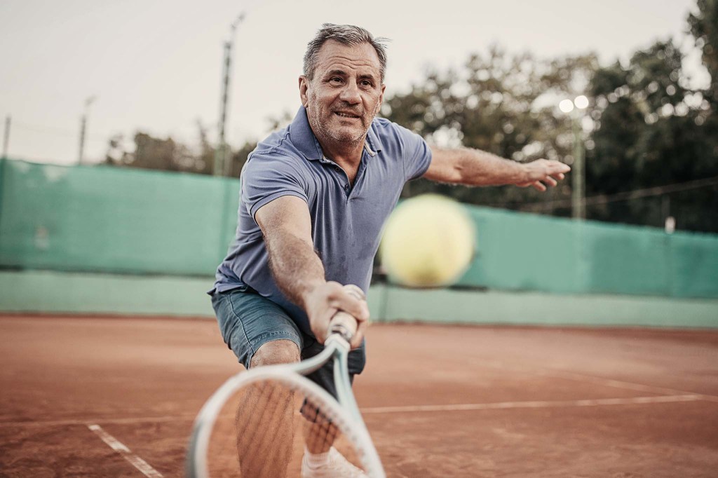 Ein älterer Mann spielt Tennis. Er trägt dunkelblaue Shorts und ein blaues Hemd und schlägt gerade den Ball übers Netz.