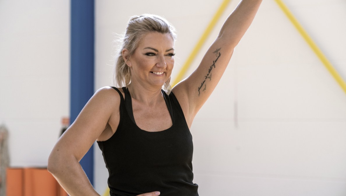 Moni zeigt sich in eleganter Pose in einem Tanzstudio. Ihr Haar ist blond und schulterlang. Sie trägt ein schwarzes Top. Ihren Unterarm ziert ein Tattoo. Lächelnd und mit anmutig ausgestrecktem Arm präsentiert sie sich.
