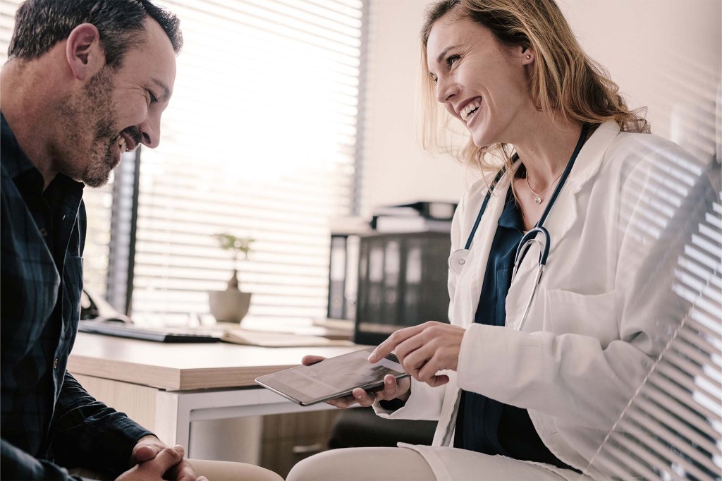 Eine blonde Ärztin entwickelt mit einem Patienten einen Ernährungsplan. Sie sitzt mit ihm in ihrer Praxis und erklärt ihm die Ernährung nach Krebs mithilfe einer Broschüre.