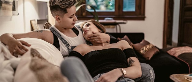 Ein junges lesbisches Paar hat es sich zuhause auf dem Sofa gemütlich gemacht. Die eine Partnerin liegt auf dem Schoß der anderen. Die beiden sehen sich verliebt an. Die sitzende Partnerin hat ihren Arm um den Kopf der anderen gelegt.