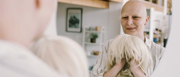 Eine Frau, die ihre Haare während der Krebstherapie verloren hat, steht vor einem Spiegel. In ihren Händen hält sie eine blonde Langhaarperücke.