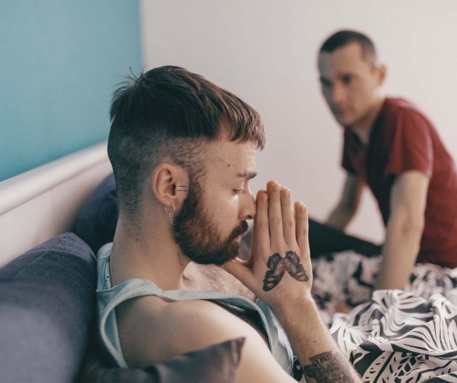 Ein schwules Paar sitzt auf dem gemeinsamen Bett. Die Stimmung wirkt angespannt. Ein Partner hält seine Hände zusammengefaltet vor dem Gesicht und die Augen geschlossen, als würde er seine Gedanken sammeln, der andere schaut ihn aus einigem Abstand an.