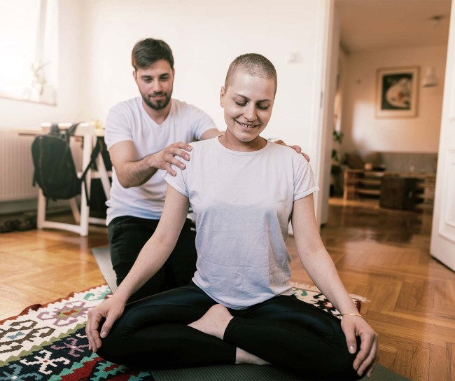 Eine junge Frau, die eine Krebsbehandlung überstanden hat, hockt im Schneidersitz auf einem Teppich im Zimmer einer Altbauwohnung. Sie trägt ein helles Shirt und schwarze Hosen und hat die Hände auf ihre Knie gelegt. Hinter ihr sitzt ihr Partner, ebenfalls im hellen T-Shirt und schwarzen Trainingshosen. Er hat seine Hände auf ihre Schultern gelegt und stützt ihre Haltung.