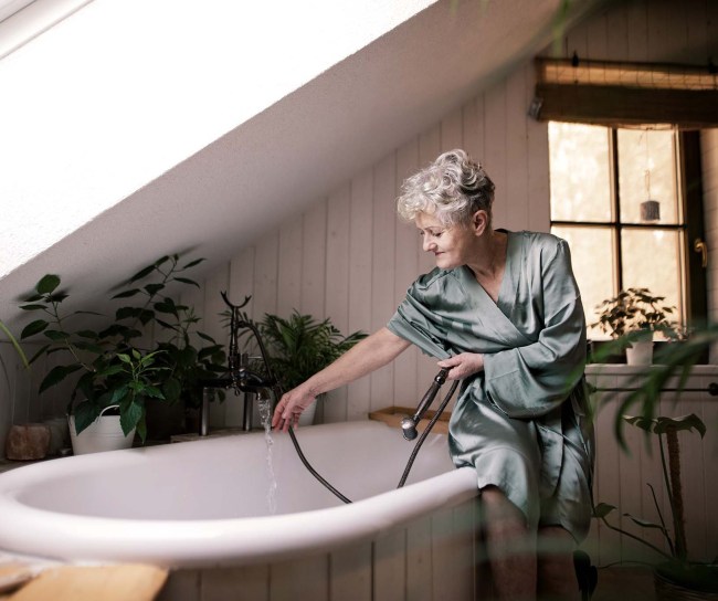 Eine ältere Frau sitzt auf ihrem Badewannenrand, während sie das Wasser in der Badewanne einlässt. Viele grüne Pflanzen schenken dem Badezimmer eine angenehme Atmosphäre.