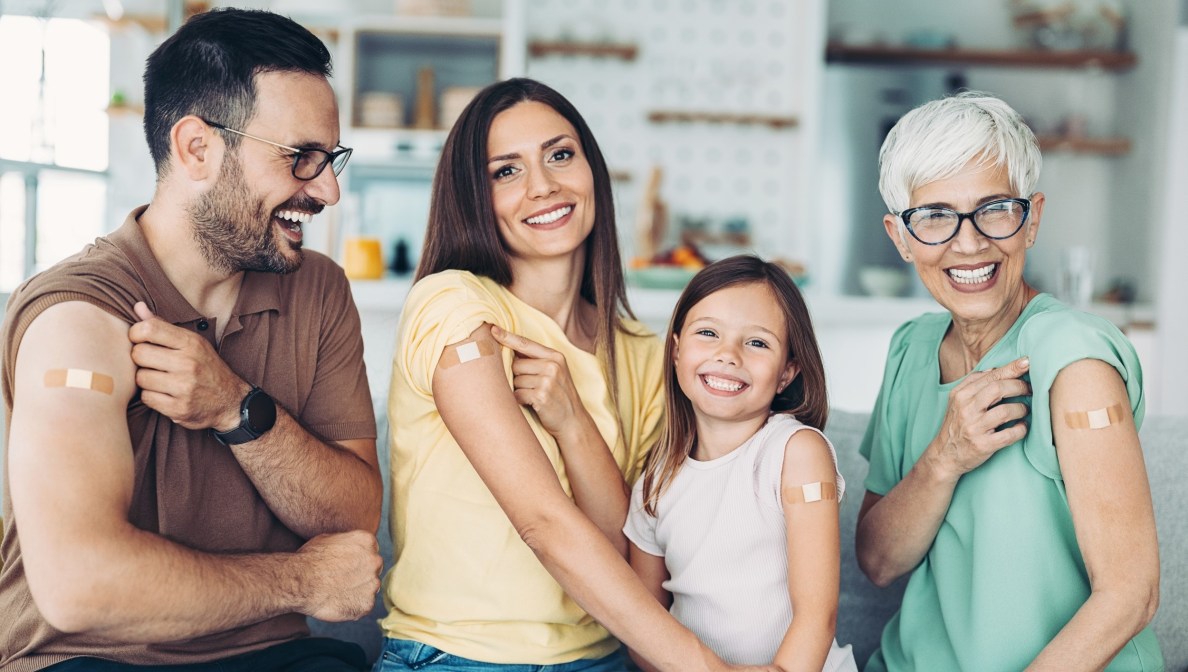 Eine Familie, Eltern, Tochter und Großmutter, blicken lächelnd in die Kamera. Alle haben einen Arm zur Kamera gedreht und haben ein kleines Pflaster auf dem Oberarm.