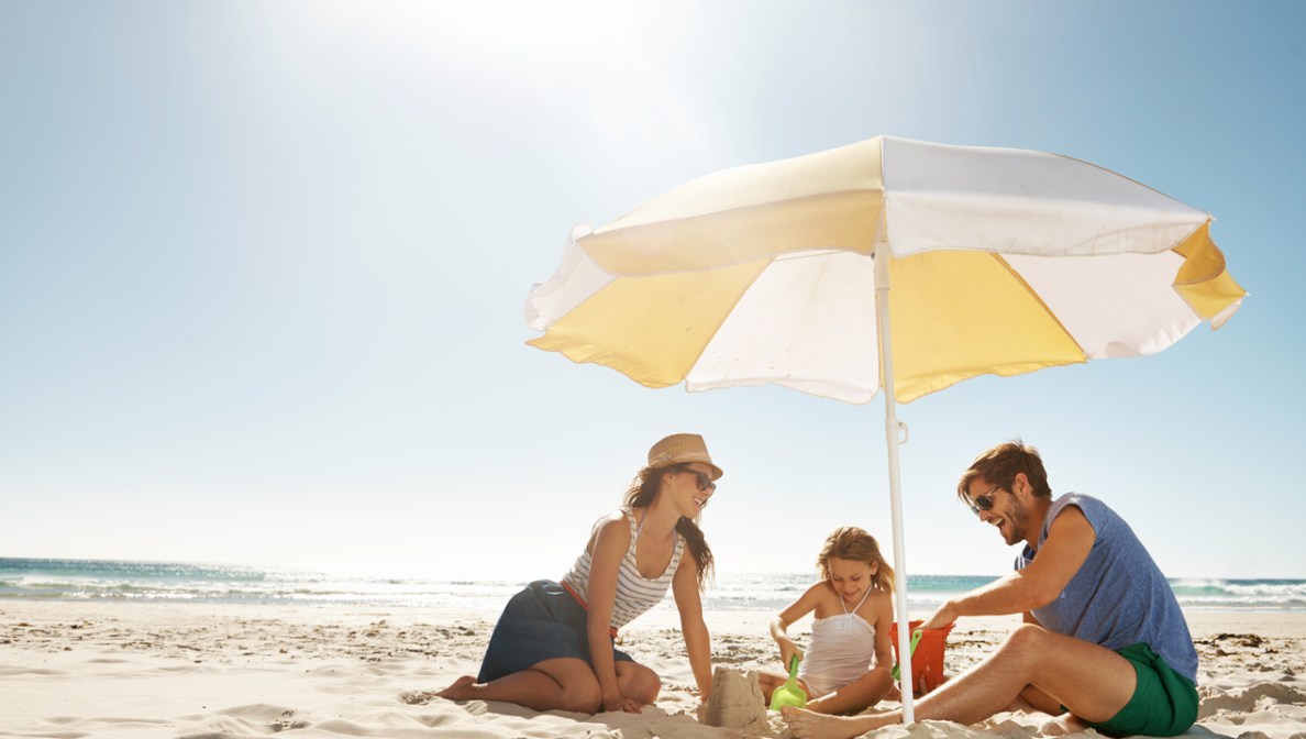 Eine Familie sitzt am Strand und spielt im Sand, im Hintergrund ist Meer zu sehen. Sie sitzen unter einem Sonnenschirm. Die Eltern tragen Sommerkleidung, das Kind einen Badeanzug.