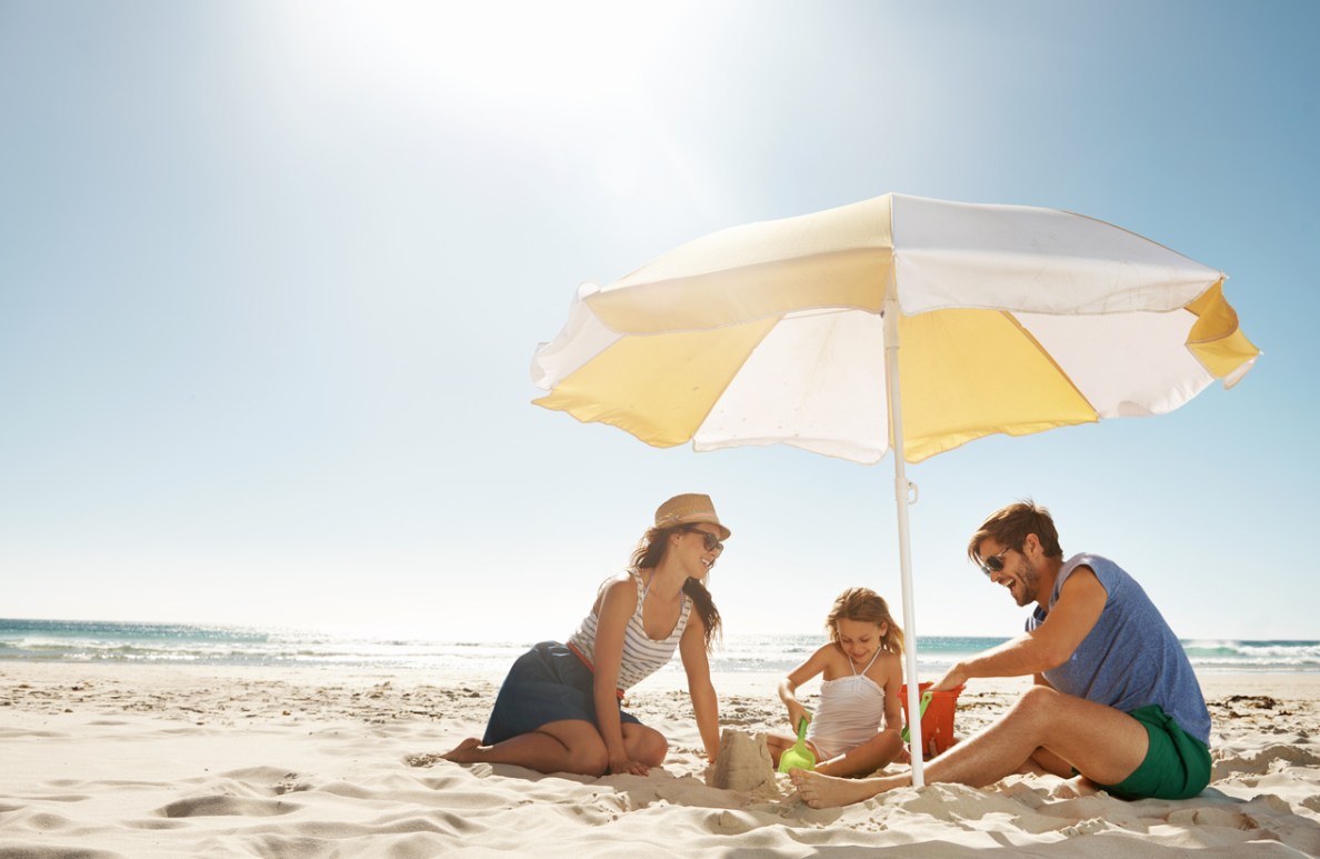 Eine Familie sitzt am Strand und spielt im Sand, im Hintergrund ist Meer zu sehen. Sie sitzen unter einem Sonnenschirm. Die Eltern tragen Sommerkleidung, das Kind einen Badeanzug.
