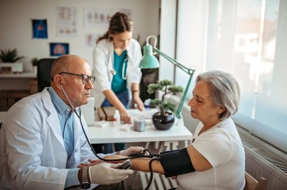 Ein Arzt misst im Sprechzimmer den Blutdruck einer älteren Patientin. Im Hintergrund ist eine Arzthelferin zu sehen, die Unterlagen auf dem Tisch ansieht.