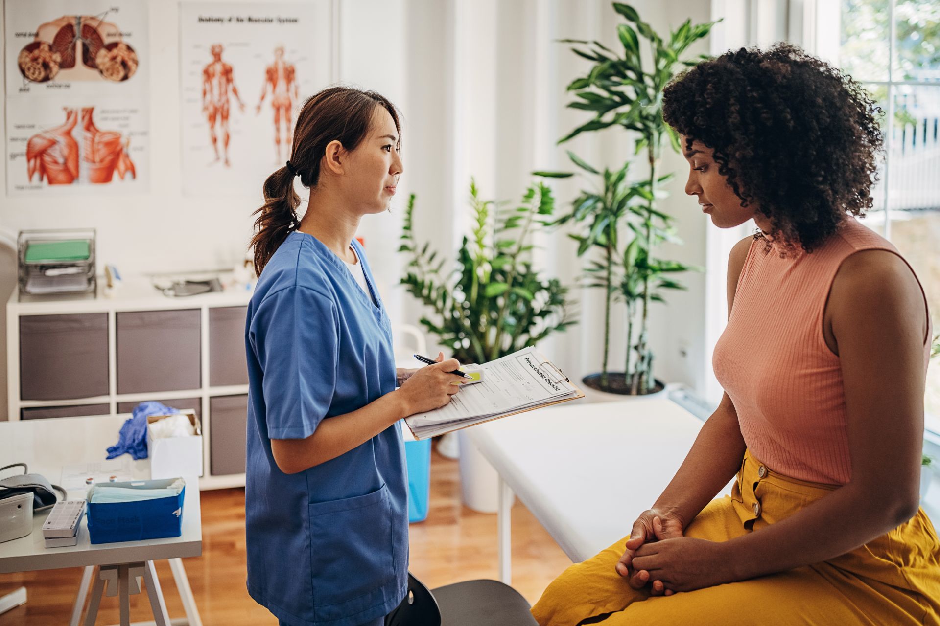 Eine Patientin sitzt auf einer Behandlungsliege beim Arzt. Die Ärztin steht vor ihr mit einem Klemmbrett und einem Stift in der Hand. Auf dem Klemmbrett ist ein Formular zu sehen.