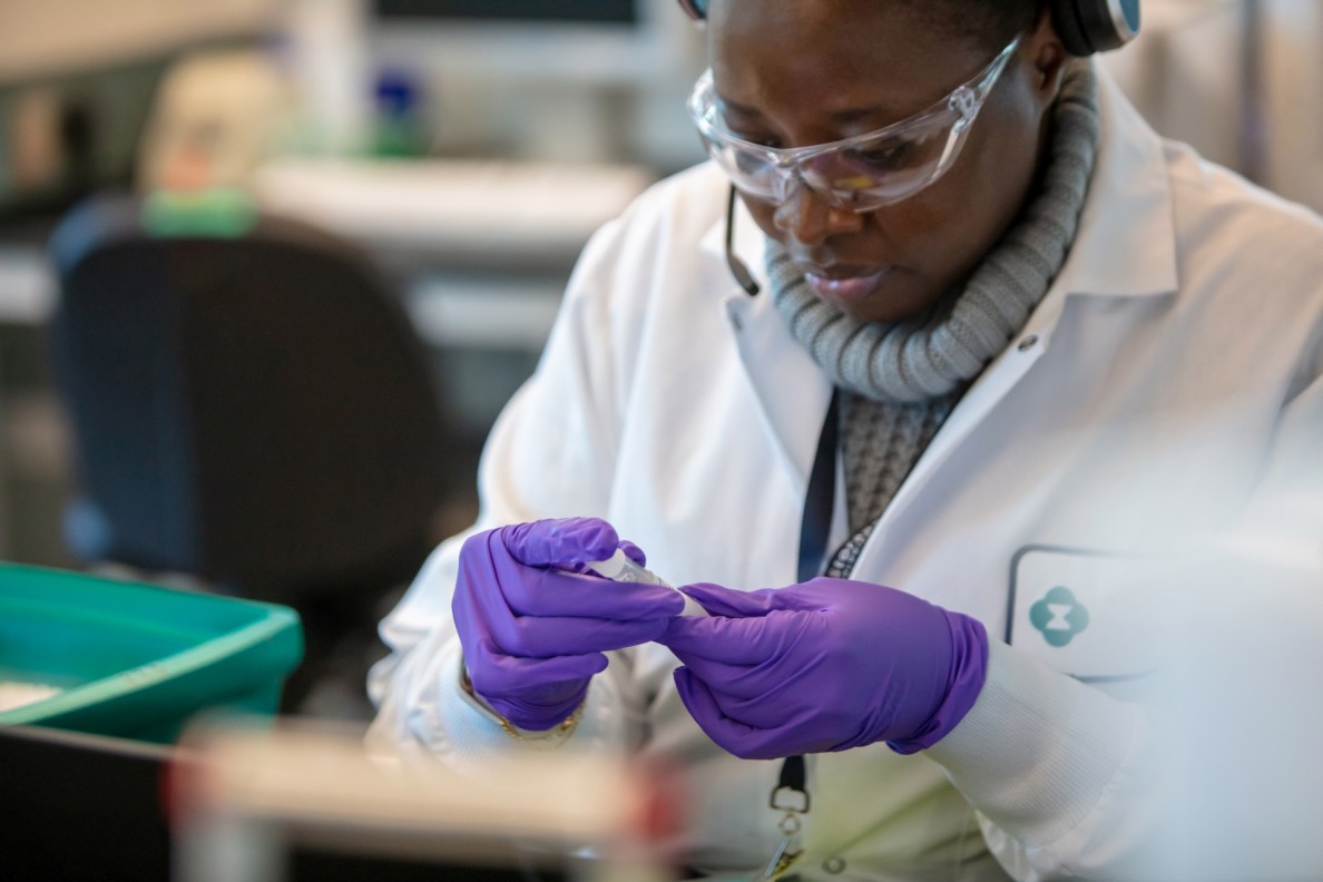 Eine Frau steht im Labor und trägt Kittel, Gummihandschuhe und Schutzbrille. Sie betrachtet eingehend ein kleines Plastikgefäß, das sie in der Hand hält.