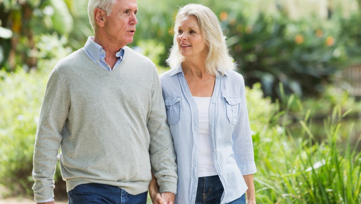 Ein Mann und eine Frau laufen Hand in Hand. Der Mann spricht, die Frau schaut ihn dabei an. Im Hintergrund sind Bäume und Sträucher zu sehen.