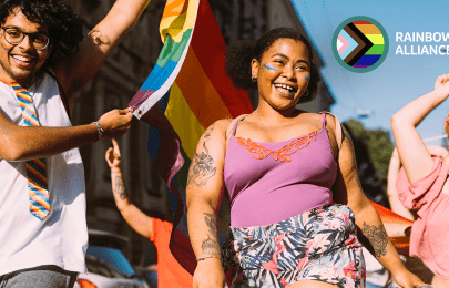 Das Bild zeigt tanzende Menschen bei einer Parade. Im Vordergrund schwingt ein Mann eine Regenbogenfahne und trägt eine Regenbogenkrawatte. Eine Frau hat Regenbogen auf die Wangen gemalt. In der oberen rechten Ecke des Bildes ist das Logo der MSD Rainbow Alliance eingefügt.