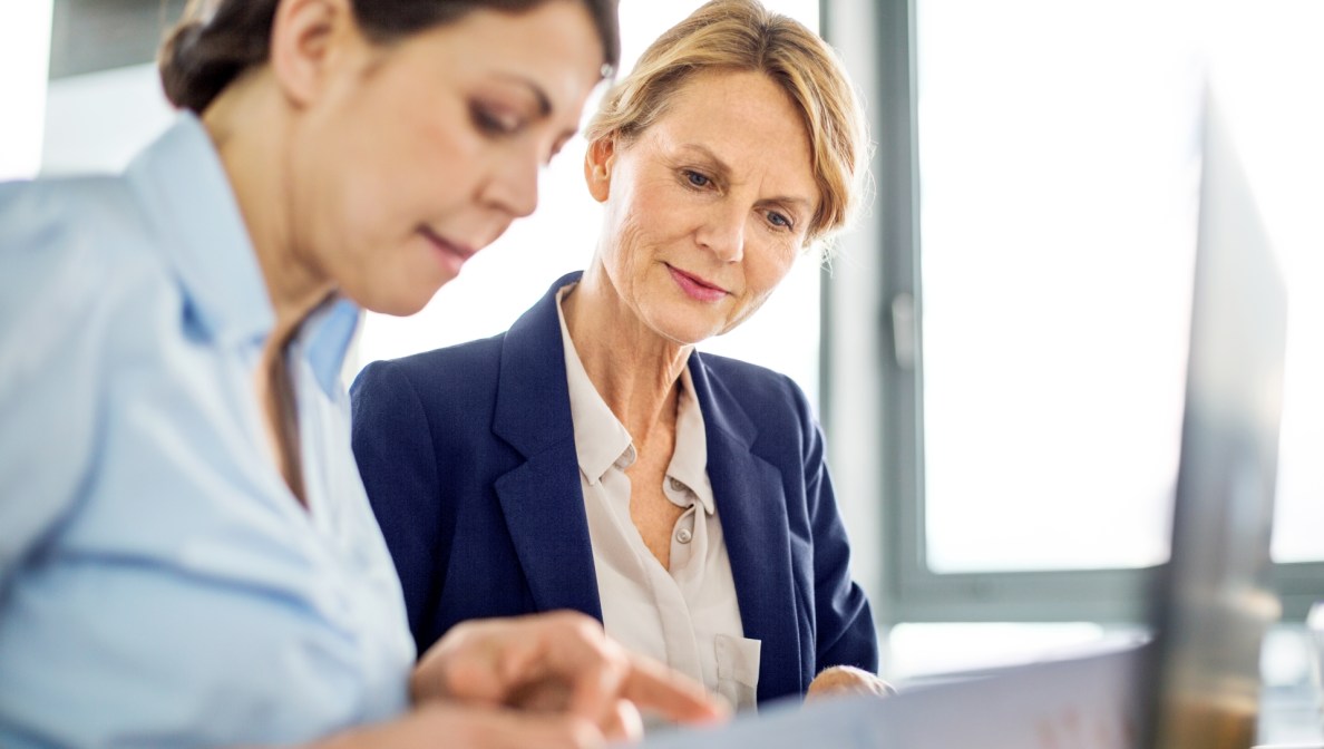 Zwei Frauen sitzen nebeneinander und blicken gemeinsam auf etwas, das außerhalb des Blickfelds des Betrachters liegt. Im Vordergrund ist ein aufgekappter Laptop zu sehen.