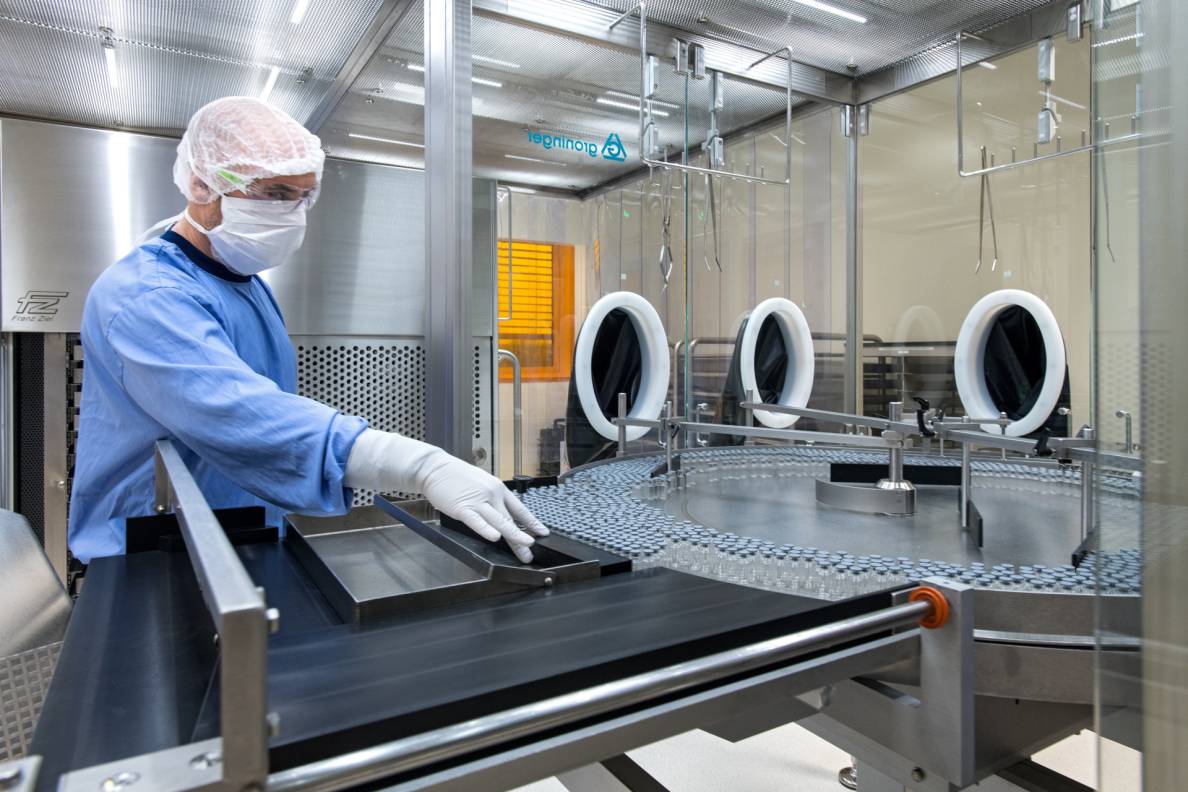 Eine Person in Hygienekleidung steht hinter einer Maschine, in die Glasfläschchen zur Abfüllung des Impfstoffs laufen.