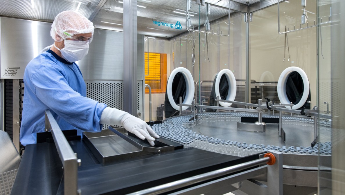 Eine Person in Hygienekleidung steht hinter einer Maschine, in die Glasfläschchen zur Abfüllung des Impfstoffs laufen.