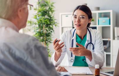 Eine junge Ärztin erklärt einem Patienten etwas. Die Ärztin ist mit dem Gesicht der Kamera zugewandt. Den Patienten sieht man lediglich seitlich von hinten, sodass nur ein Teil des Rückens und des Gesichts zu sehen ist.
