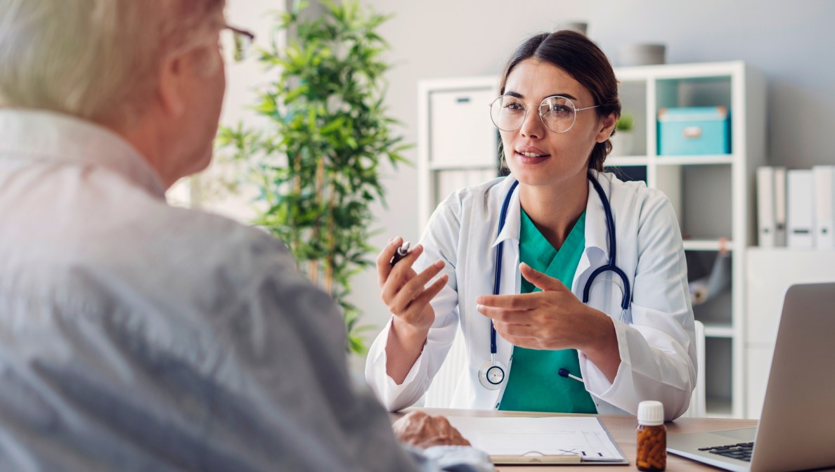 Eine junge Ärztin erklärt einem Patienten etwas. Die Ärztin ist mit dem Gesicht der Kamera zugewandt. Den Patienten sieht man lediglich seitlich von hinten, sodass nur ein Teil des Rückens und des Gesichts zu sehen ist.