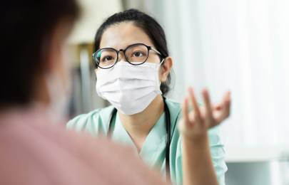 Eine Ärztin mit Mundschutz sitzt in Praxisräumen und erklärt etwas. Im Vordergrund des Bildes ist ein Patient im Anschnitt von hinten zu sehen.
