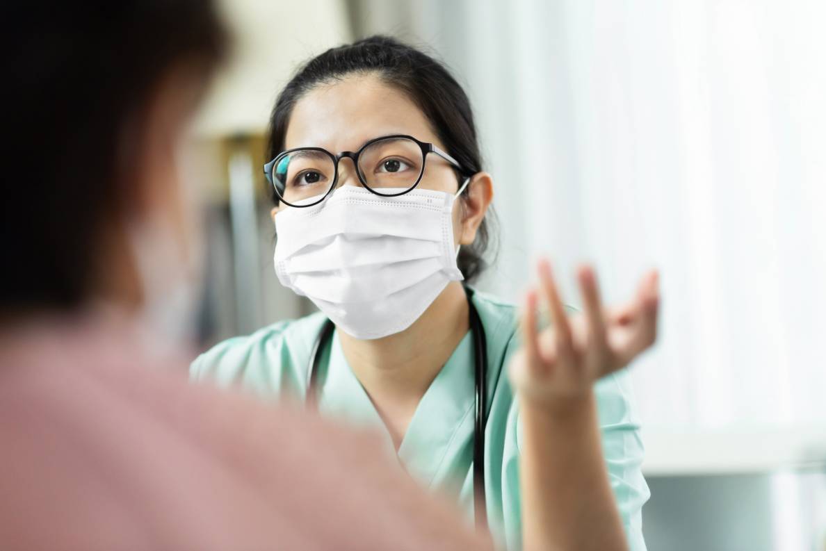 Eine Ärztin mit Mundschutz sitzt in Praxisräumen und erklärt etwas. Im Vordergrund des Bildes ist ein Patient im Anschnitt von hinten zu sehen.