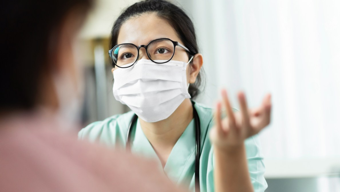 Eine Ärztin mit Mundschutz sitzt in Praxisräumen und erklärt etwas. Im Vordergrund des Bildes ist ein Patient im Anschnitt von hinten zu sehen.
