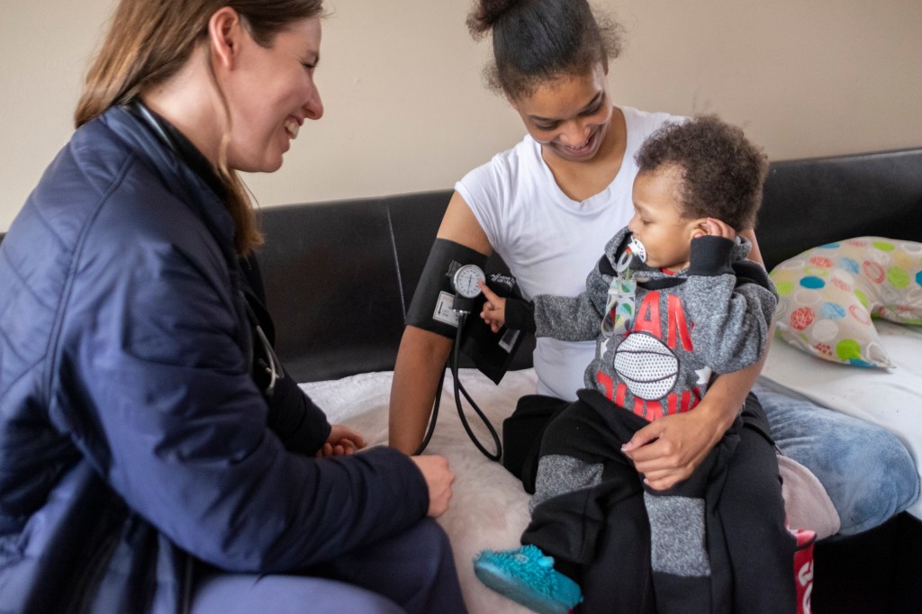 Zwei Frauen sitzen auf dem Sofa. Die rechte Frau hat eine Blutdruckmanschette um den rechten Arm und ein Kind auf dem Schoß.