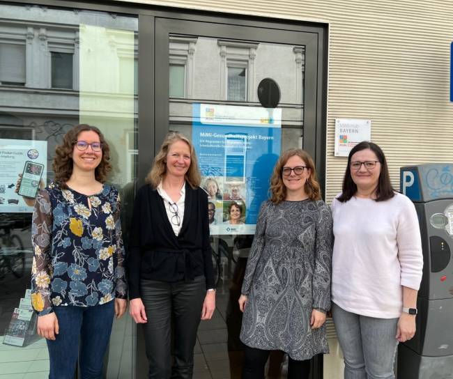 Vier Frauen stehen vor einem Plakat zum MiMi-Gesundheitsprojekt Bayern