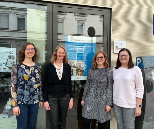 Vier Frauen stehen vor einem Plakat zum MiMi-Gesundheitsprojekt Bayern