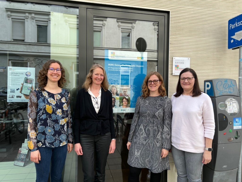 Vier Frauen stehen vor einem Plakat zum MiMi-Gesundheitsprojekt Bayern