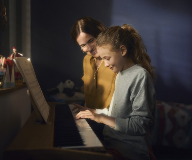 Ein junges Mädchen spielt Klavier. Die Mutter sitzt neben ihr und schaut ihr dabei zu.