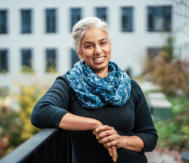 Eine Frau mit grauen Haaren steht lächelnd im Freien an einem Geländer. Sie trägt einen dunkelblauen Pullover und einen gemusterten blauen Schal. Im Hintergrund ist ein modernes Gebäude mit vielen Fenstern sowie herbstlich gefärbte Pflanzen zu sehen. Die Atmosphäre wirkt freundlich und entspannt.