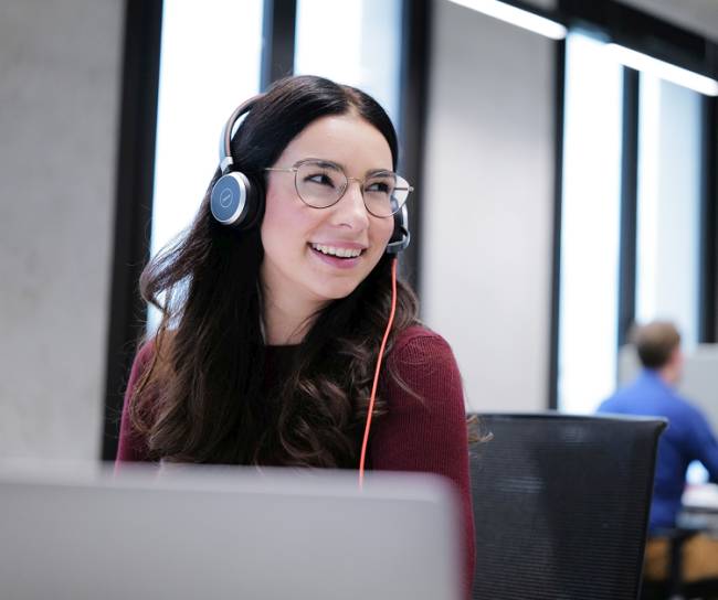 Eine junge Frau mit Brille und Headset sitzt an einem Schreibtisch in einem Großraumbüro und lächelt. Vor ihr steht ein Laptop, sie wirkt freundlich und engagiert im Kundengespräch.