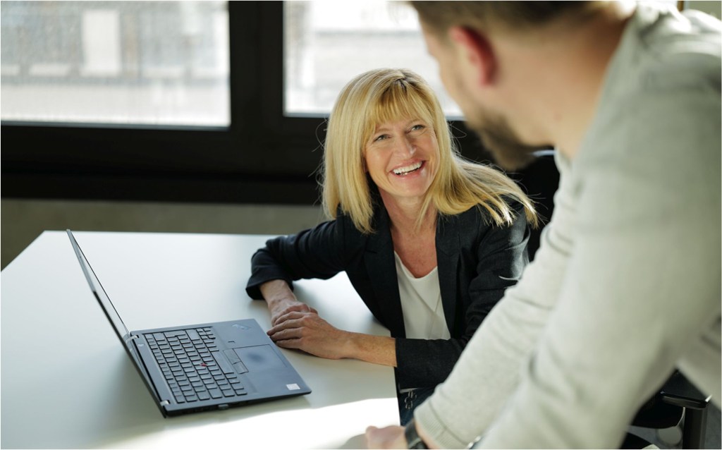 Eine blonde Frau in Businesskleidung sitzt an einem Tisch mit einem aufgeklappten Laptop und lächelt einen männlichen Kollegen an, der ihr gegenübersteht. Die Szene findet in einem hellen Büro mit großen Fenstern statt und wirkt offen und sympathisch.