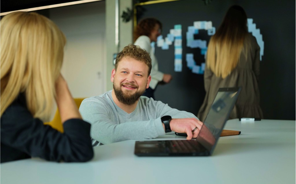 Ein Mann mit lockigem Haar und Bart sitzt an einem Tisch in einem modernen Büro und spricht lächelnd mit einer Frau, die ihm gegenüber sitzt. Er zeigt auf seinen Laptop. Im Hintergrund arbeiten zwei weitere Frauen an einer schwarzen Wand, die mit bunten Post-its in der Form von Buchstaben oder Zahlen (u. a. „MS“) beklebt ist. Die Szene wirkt lebendig und kollaborativ.