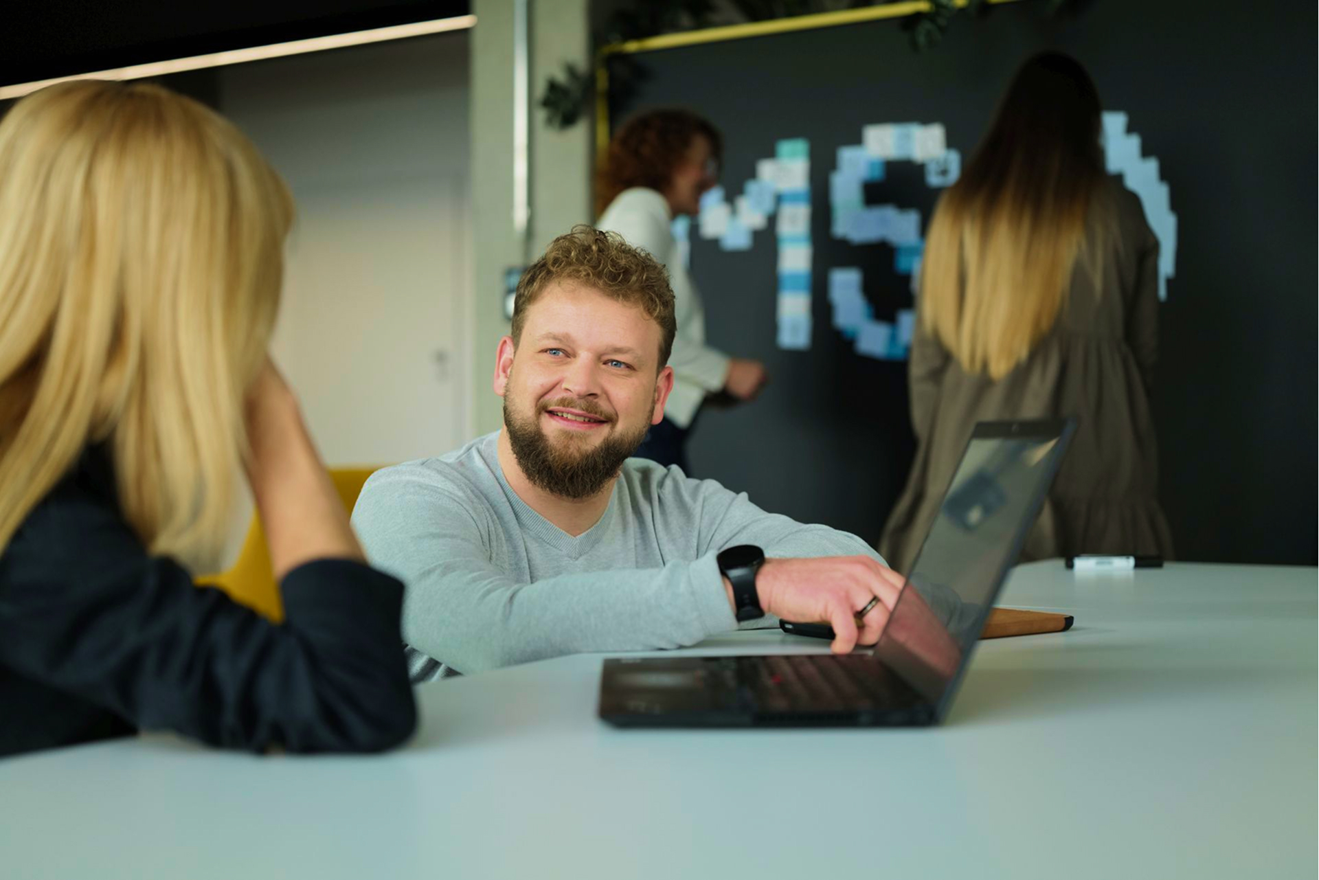Ein junger Mann mit kurzen Haaren und Bart steht an einem Stehschreibtisch in einem modernen Büro. Er trägt ein helles Poloshirt, telefoniert mit einem Smartphone und arbeitet gleichzeitig an einem Laptop. Durch eine Glaswand sind unscharf weitere Personen im Hintergrund erkennbar.