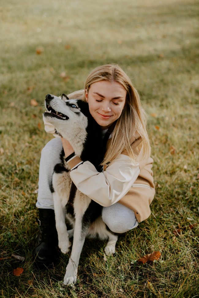 Eine junge Frau mit blonden Haaren sitzt auf einer Wiese und umarmt liebevoll einen Husky mit blauen Augen. Sie hat die Augen geschlossen und lächelt zufrieden, während der Hund den Kopf leicht hebt. Die Szene strahlt Nähe, Vertrauen und Harmonie zwischen Mensch und Tier aus.