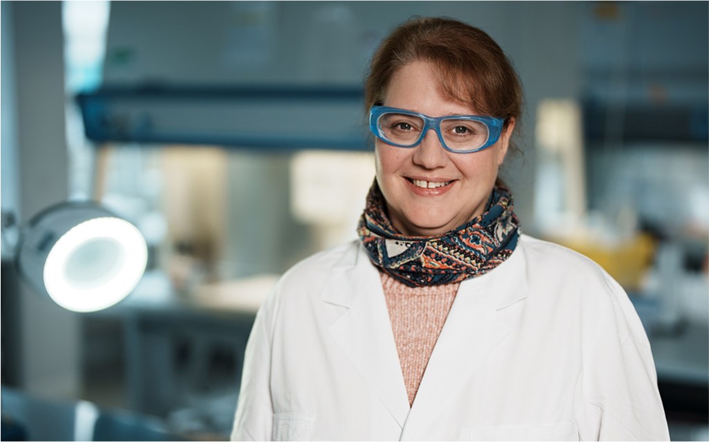 Eine Frau in einem Labor trägt einen weißen Laborkittel, eine bunte gemusterte Halsschleife und eine blaue Schutzbrille. Sie lächelt in die Kamera. Im Hintergrund ist ein Laborarbeitsplatz mit Beleuchtung und technischen Geräten zu sehen. Die Atmosphäre wirkt professionell und freundlich.