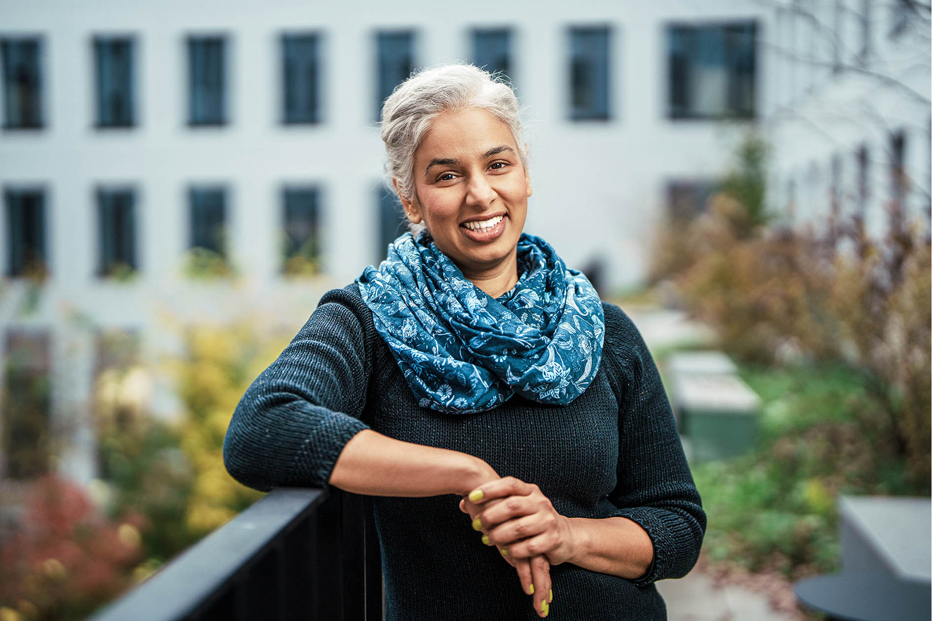Eine Frau mit grauen Haaren steht lächelnd im Freien an einem Geländer. Sie trägt einen dunkelblauen Pullover und einen gemusterten blauen Schal. Im Hintergrund ist ein modernes Gebäude mit vielen Fenstern sowie herbstlich gefärbte Pflanzen zu sehen. Die Atmosphäre wirkt freundlich und entspannt.