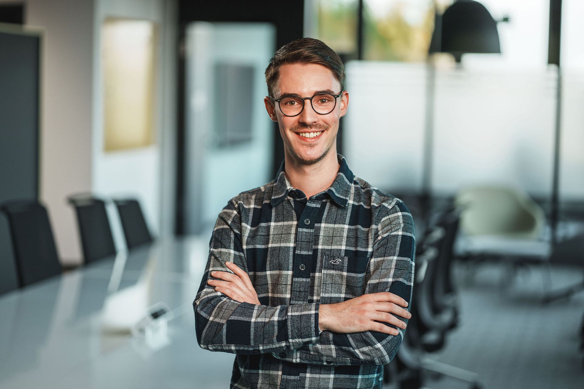 Ein junger Mann mit Brille und kariertem Hemd steht lächelnd mit verschränkten Armen in einem modernen Konferenzraum. Der Hintergrund zeigt einen Besprechungstisch und Glaswände, die eine professionelle, aber freundliche Atmosphäre vermitteln.