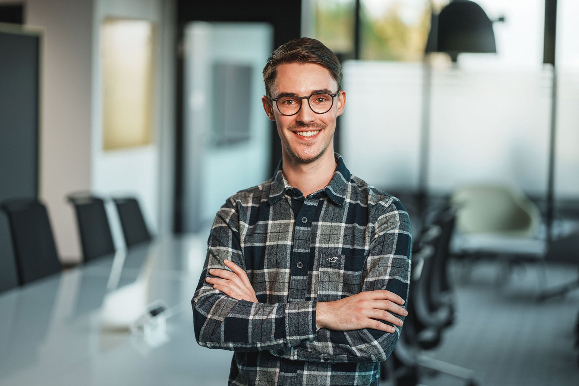 Ein junger Mann mit Brille und kariertem Hemd steht lächelnd mit verschränkten Armen in einem modernen Konferenzraum. Der Hintergrund zeigt einen Besprechungstisch und Glaswände, die eine professionelle, aber freundliche Atmosphäre vermitteln.